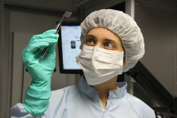 Scientist in a cleanroom suit examining a small chip with tweezers in a laboratory setting, with lab equipment and a computer screen in the background.