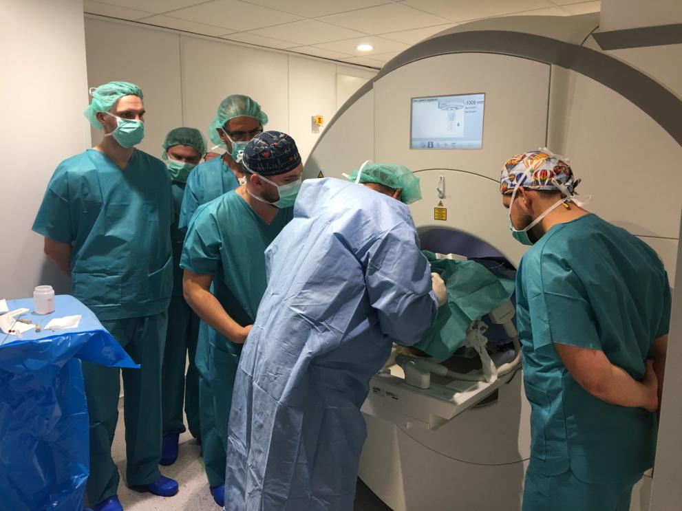 Group of surgeons in green scrubs and surgical caps gathered around a patient undergoing a procedure with a large medical imaging machine, demonstrating the precision and teamwork involved in gene therapy for Parkinson’s disease.