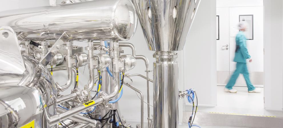 Close-up of a stainless steel spray drying machine with complex piping and valves in a clean, white pharmaceutical lab, with a blurred operator in protective gear walking in the background.