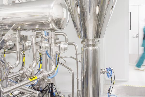 Close-up of a stainless steel spray drying machine with complex piping and valves in a clean, white pharmaceutical lab, with a blurred operator in protective gear walking in the background.